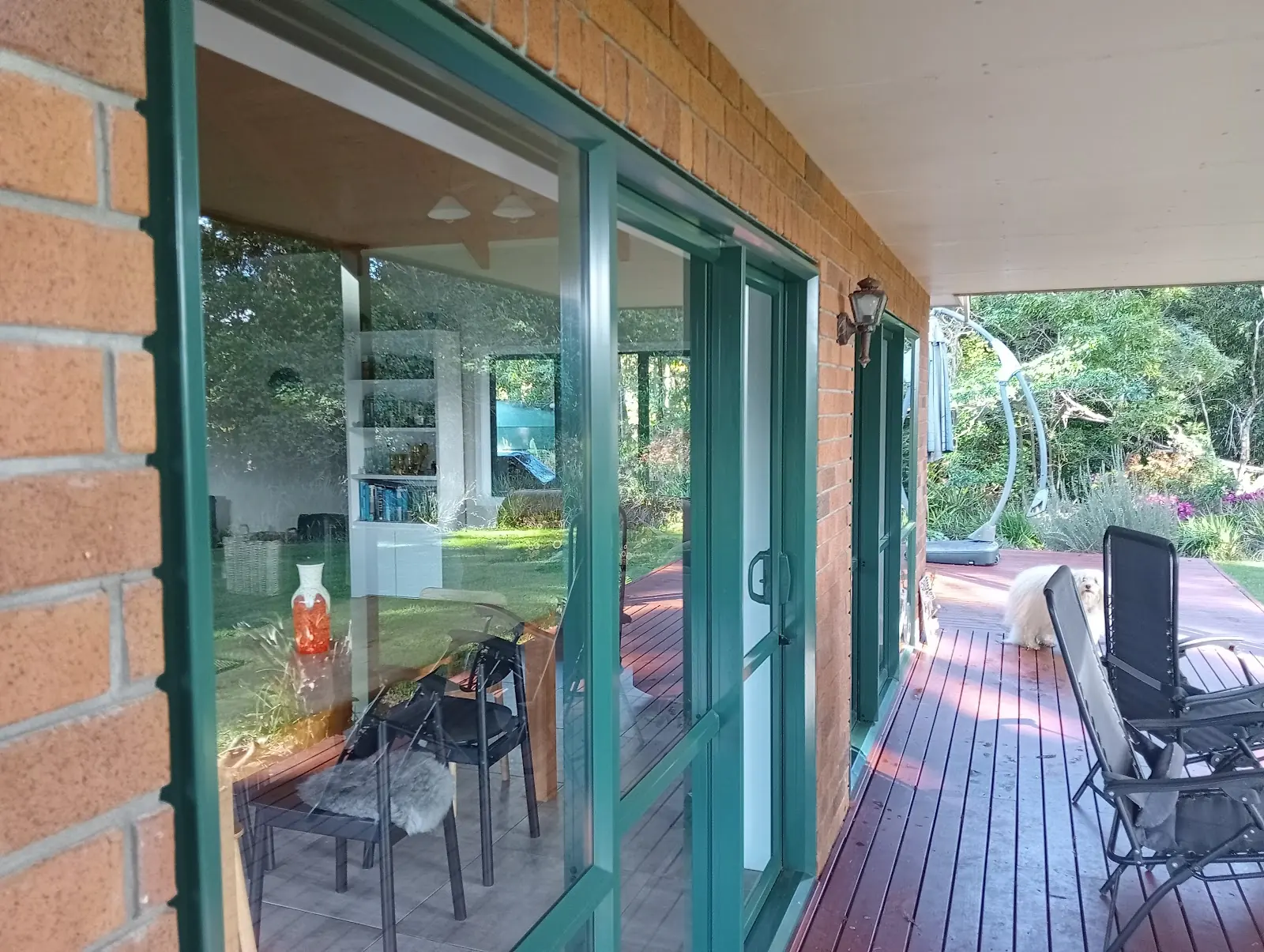 Green framed glass doors along a brick house and timber deck, with a dog lying further along
