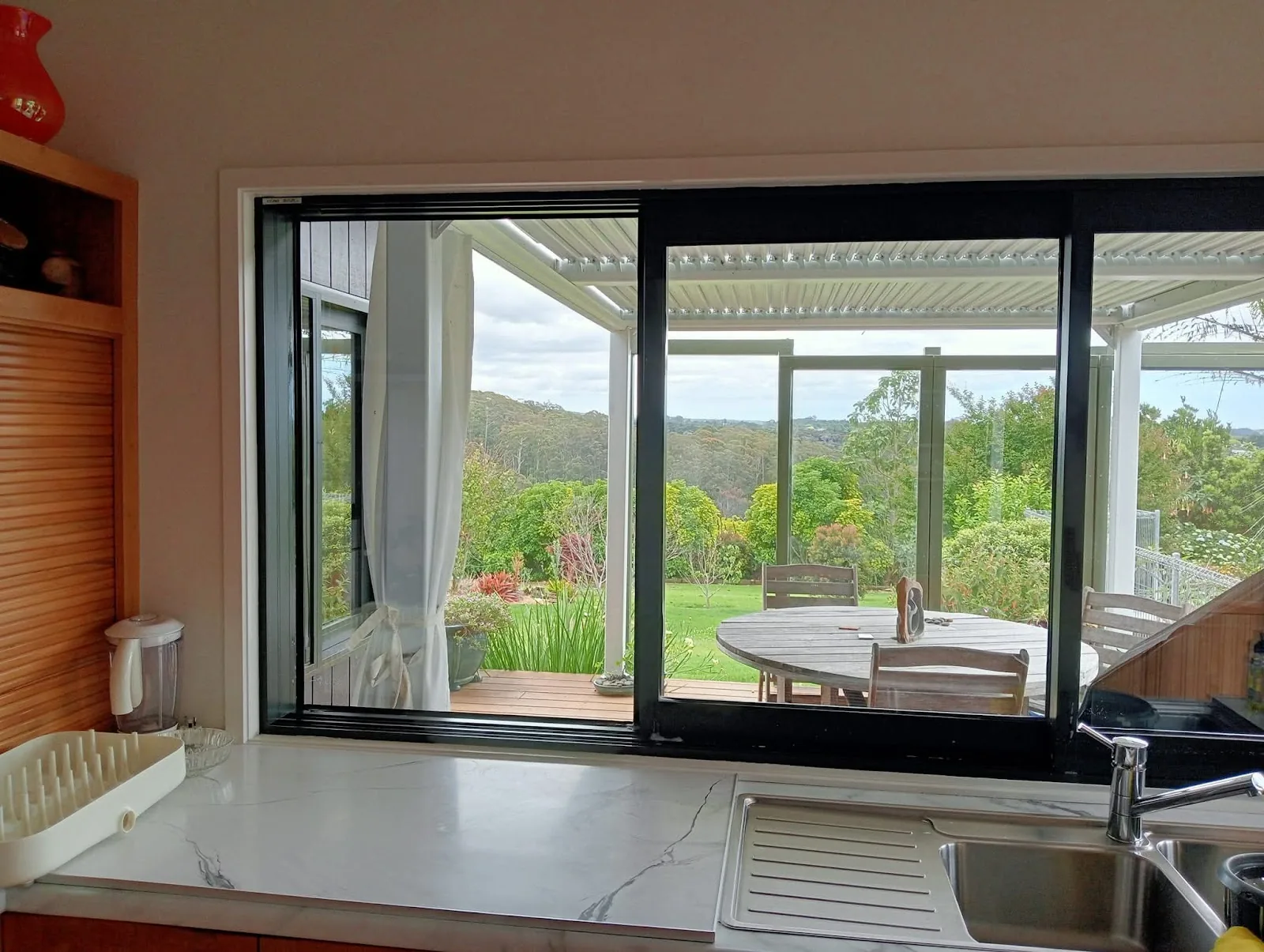 A kitchen bench looking out through a wide sliding door to a covered deck, table and garden